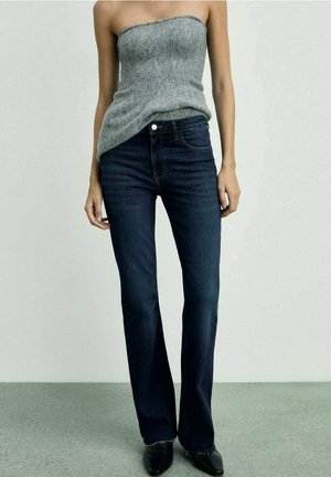 Kobieta ubrana w szary sweter bez ramiączek, ciemnoniebieskie wysoki stan jeansy oraz czarne spiczaste buty stoi na tle jednolitego tła.