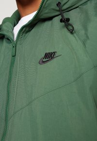 Green hooded jacket made of lightweight fabric, featuring a front zipper, adjustable drawstrings, and a black Nike logo on the chest.