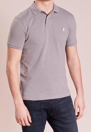 Mężczyzna w dopasowanej szarej krótkiej koszulce polo z małym białym logo konia, połączonej z ciemnoniebieskimi jeansami.