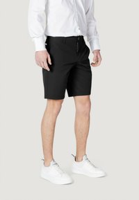 Pantalones cortos negros con un diseño a medida, que presentan un frente plano, bolsillos laterales y un cierre con botón. Llevados con zapatillas blancas y una camiseta blanca.