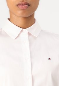 Lichtroze overhemd met kraag en verticale strepen, voorzien van knoopsluiting en een klein geborduurd logo aan de linkerkant. Glad textuur.
