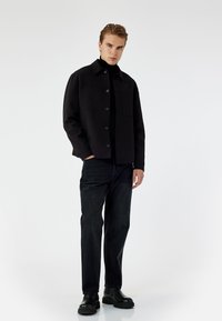 Chaqueta negra con dos bolsillos frontales, cierre de botones y un cuello clásico. Combinada con jeans de mezclilla oscura y botas negras robustas.