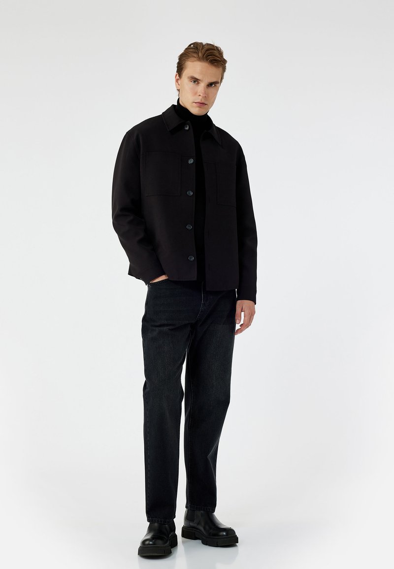 Chaqueta negra con dos bolsillos frontales, cierre de botones y un cuello clásico. Combinada con jeans de mezclilla oscura y botas negras robustas.