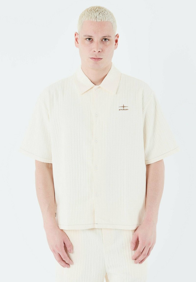 Camisa de botones de manga corta en color crema claro con una sutil textura vertical, cuello puntiagudo y un pequeño logotipo bordado en el pecho.