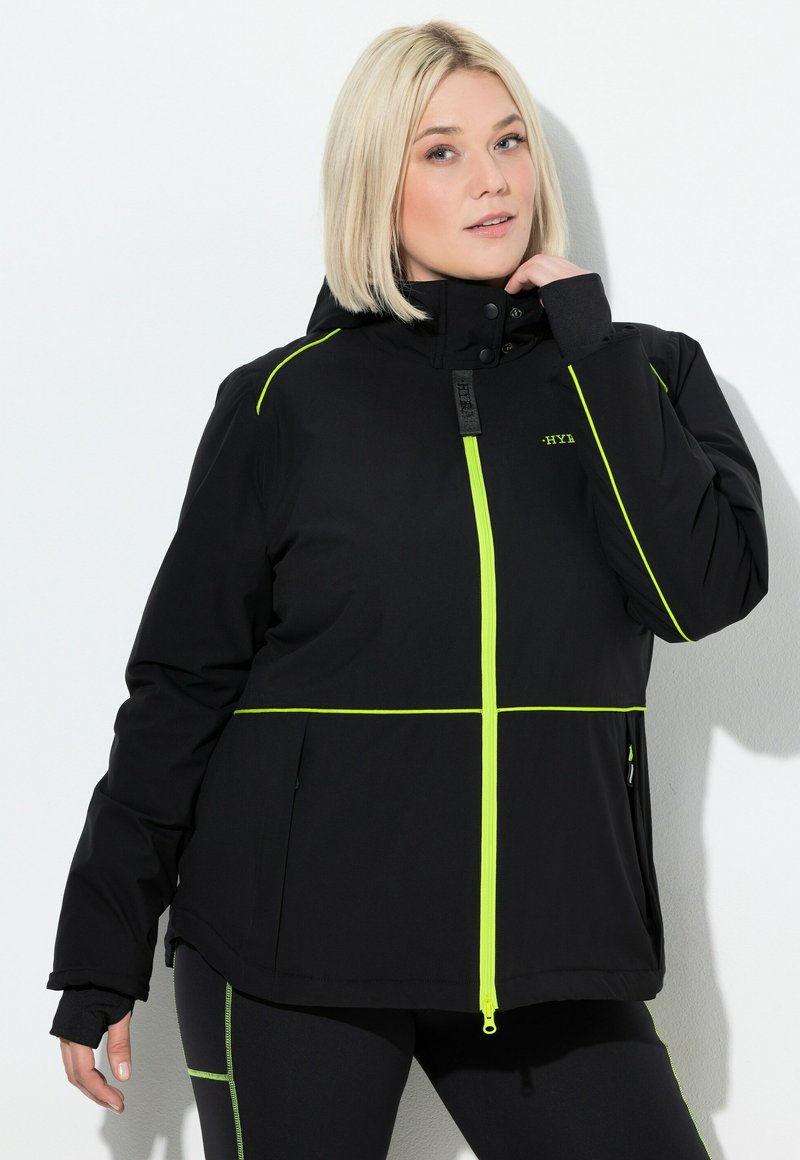 Blondýnka v černé bundě s neonově žlutým zipem a lemováním pózuje se jednou rukou u krku před jednobarevným bílým pozadím.