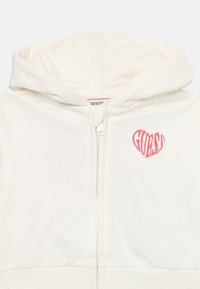White zip-up hoodie made of soft fabric with a pink "GUESS" heart logo on the chest. Features a drawstring hood and ribbed cuffs.