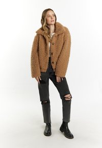 Brun faux fur-jakke med stor krave og knaplukning, parret med slidte sorte jeans og sorte ankelstøvler, sat mod en hvid baggrund.