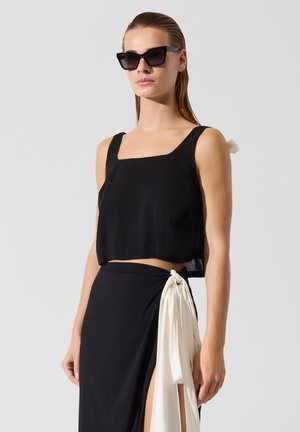 Femeie care poartă un top negru fără mâneci cu gât pătrat și o fustă neagră tip petrecut cu legătură albă pe lateral, asortată cu ochelari de soare negri cu rame cat-eye.