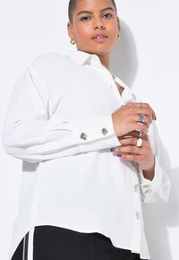 Modelo vistiendo una camisa blanca de botones con botones decorativos en los puños, exhibiendo accesorios en ambas manos y un maquillaje de ojos brillante, contra un fondo claro.