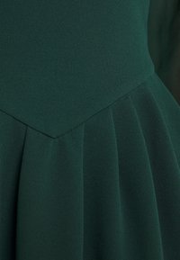 Mörkgrön klänning, tillverkad av strukturerat material. Har en plisserad kjol med en V-formad bodice och släta sömmar.