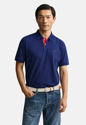 Man draagt een marineblauw poloshirt met een rode binnenkant van de knoopsluiting, een witte geweven riem en blauwe spijkerbroek, staand met één hand in de broekzak.