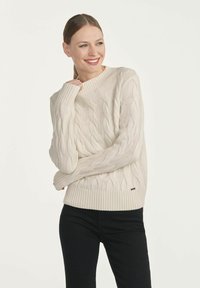 Sweter w kolorze kremowym, wykonany z splotu warkoczowego, z prążkowanym okrągłym dekoltem i mankietami. Na dole znajduje się mała metka z logo. Noszony z czarnymi spodniami.