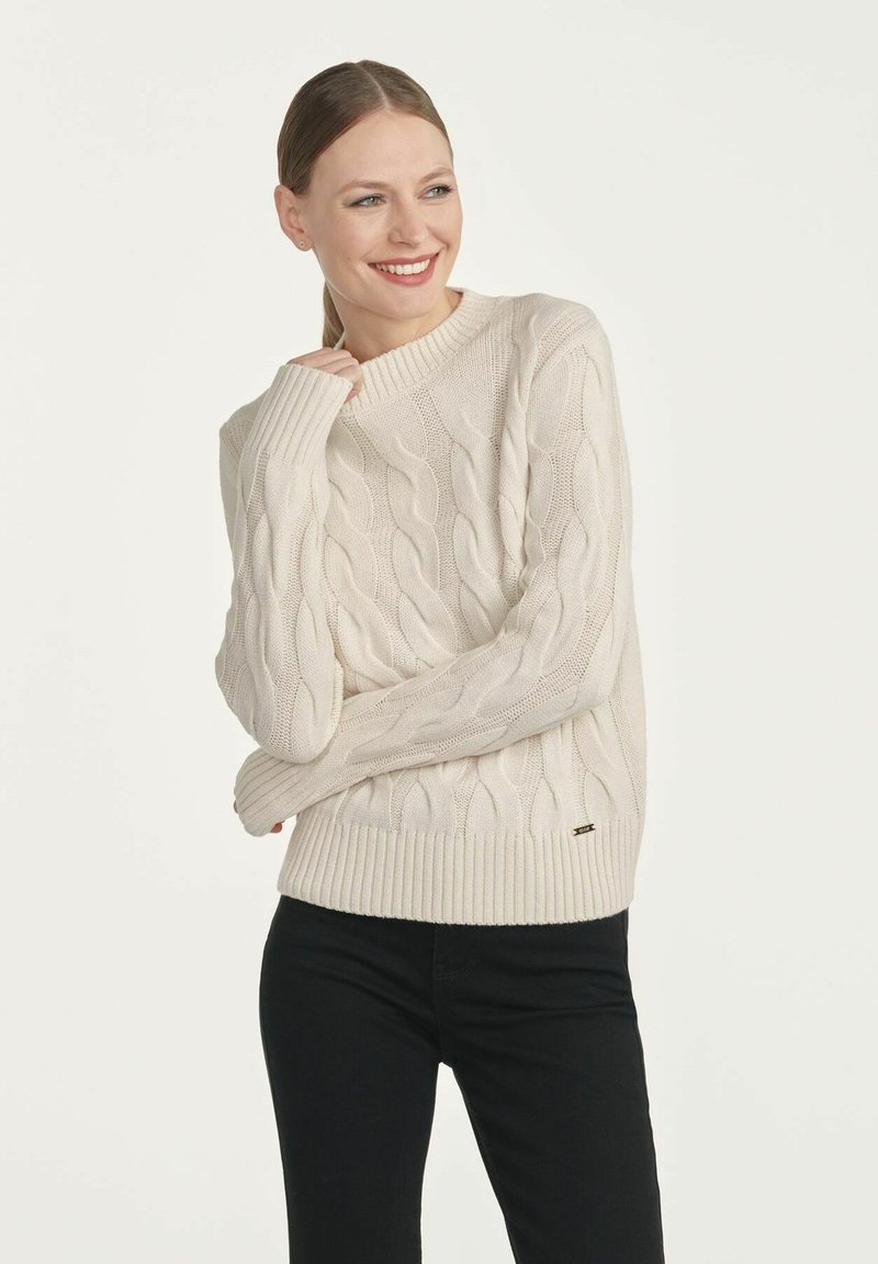 Sweter w kolorze kremowym, wykonany z splotu warkoczowego, z prążkowanym okrągłym dekoltem i mankietami. Na dole znajduje się mała metka z logo. Noszony z czarnymi spodniami.