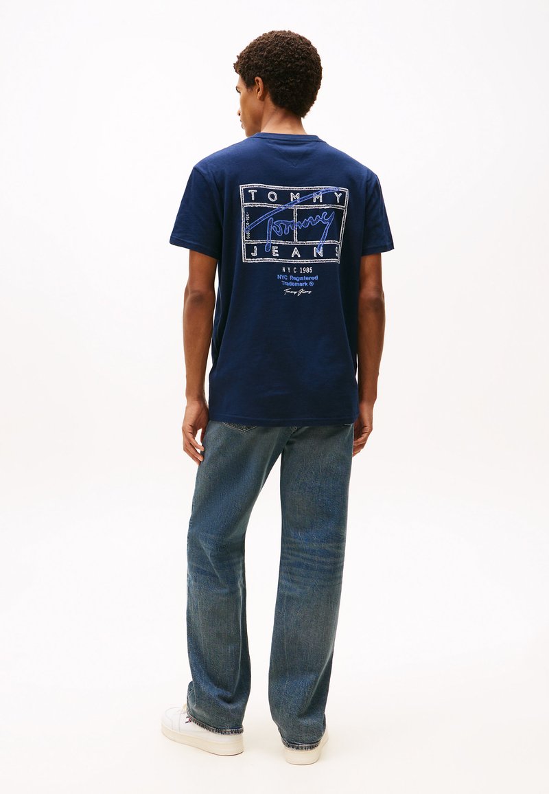 T-shirt en coton bleu marine avec un imprimé graphique blanc et bleu au dos, contrasté par un jean bleu clair et des baskets blanches.