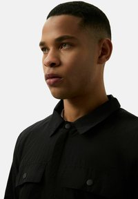 Zwarte lichte overhemd met een kraag, twee borstzakken en knoopsluiting. Gladde textuur met een subtiele glans.