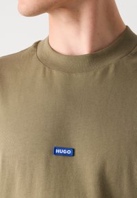 Olive roheline t-särk ribadest ümmarguse kaelusega ja väike sinine "HUGO" logo patch vasakul küljel. Pehme, siledat kangast tekstuur.