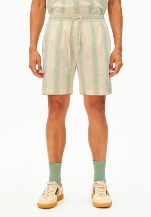 Man draagt lichtgroene en beige gestreepte shorts met trekkoord, groene sokken en beige sneakers met zwarte strepen, staand tegen een witte achtergrond.