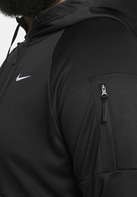 Sudadera con capucha negra de Nike, con una tela suave y texturada, que presenta un bolsillo con cremallera en la parte superior del brazo y un logo blanco en el pecho.