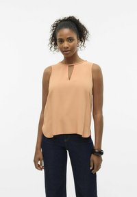 Blusa senza maniche pesca con scollatura a chiave, texture liscia e una vestibilità leggermente ampia, abbinata a jeans in denim scuro.