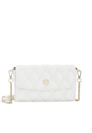 Valkoiseen mekkoon pukeutunut nainen kantaa beigeä, helmikoristeista ja kultaketjullista tikattua käsilaukkua, seisten kukkakoristeiden lähellä.