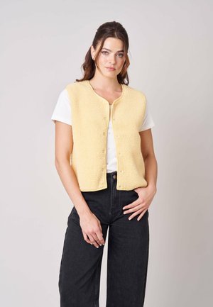 Jonge vrouw met bruin haar draagt een lichtgele mouwloze vest met knopen over een wit T-shirt en zwarte broek, staand tegen een effen achtergrond.