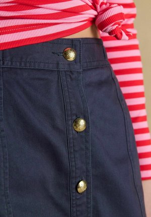 Person trägt einen marineblauen Rock mit drei goldenen Knöpfen und ein pink-rot gestreiftes Oberteil, das in der Taille verknotet ist.