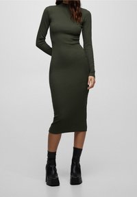 Femme debout portant une robe midi ajustée à manches longues couleur vert olive avec des bottines noires à plateforme sur un fond uni.