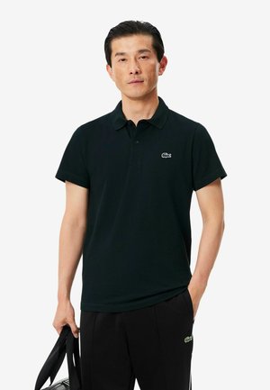 Hombre con polo negro y pantalones con pequeños logotipos de cocodrilos blancos y verdes, sosteniendo una correa negra de bolsa con una mano.