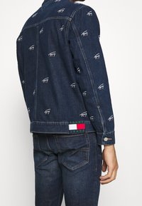 Jeansjacka i mörkblått med broderade logotyper över hela ytan. Har tryckknappar och en "TOMMY JEANS"-märke med logotyp och färgdetaljer.