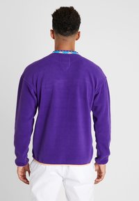 Pullover in pile viola con maniche lunghe, tessuto testurizzato, collo tondo e orlo arancione a contrasto. Presenta un colletto colorato e decorato.