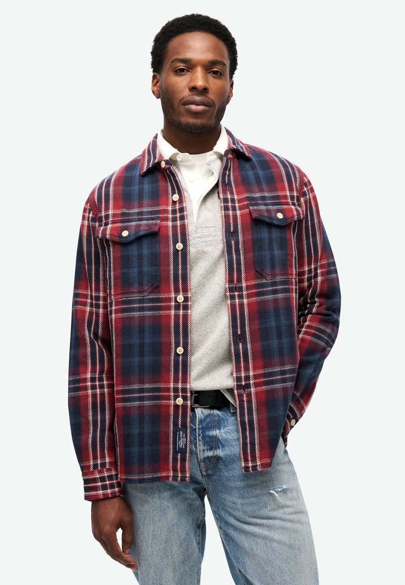 Man in een rood en marineblauw geruite overhemd, met een lichtgrijze pullover eronder en lichtblauwe spijkerbroek, staand met één hand in de broekzak.