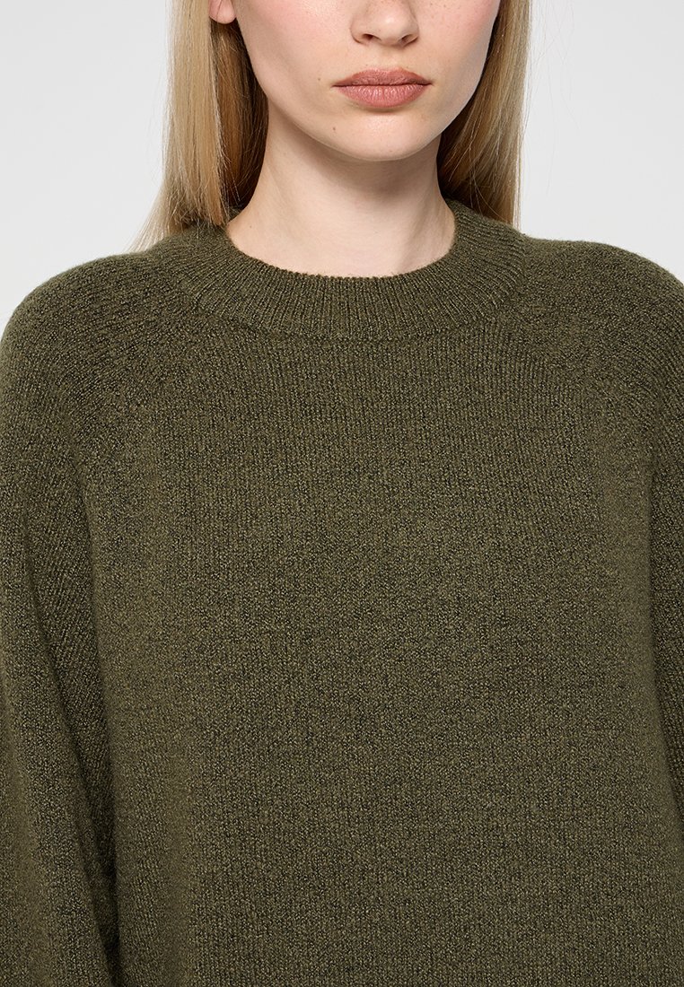 Frau trägt einen olivgrünen, gestrickten Pullover mit Rundhalsausschnitt, zeigt den Oberkörper und einen Teil ihrer glatten, blonden Haare.