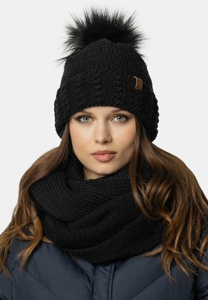 Beanie nero lavorato a maglia con un pompon in pelliccia sintetica soffice e una patch in pelle marrone chiaro. Accompagnato da una sciarpa nera lavorata a maglia spessa.