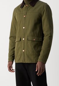 Veste vert olive avec un col en velours côtelé marron, des boutons en cuivre et deux poches avant. Fabriquée dans un tissu robuste avec une coupe décontractée.