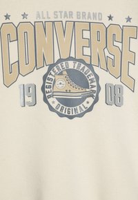Felpa beige con la scritta "CONVERSE", un logo circolare con un'illustrazione di sneakers e accenti metallici. Il testo include "ALL STAR BRAND" e delle date.