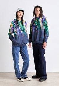 Hættetrøjer med livlige grafiske mønstre i grøn, lilla og gul, parret med blå jeans og sorte bukser. Uformelt fodtøj er inkluderet.