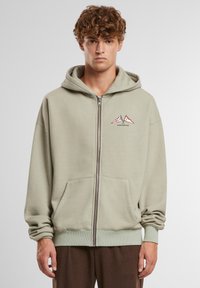 Fleeceowa bluza z kapturem w stonowanej zieleni, z regulowanym kapturem, przednimi kieszeniami i grafiką góry z napisem "ZABRONIONE". Prążkowane mankiety i dół.