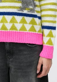 Pletený sveter s pruhovaným vzorom v zelenej, modrej a bielej farbe, s ružovými rebrovanými manžetami a trojuholníkovými motívmi na leme. Viditeľné sú sivé džínsy.