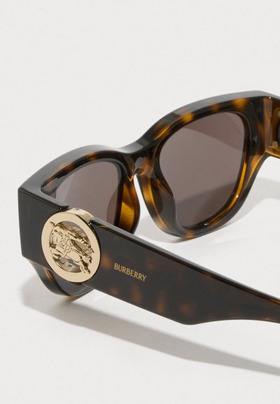 Pruun kilpkonnakujuline prilliraam tumedate läätsedega. Omab kuldset ümmargust logo käe peal. Küljel on reljeefselt kiri "BURBERRY".