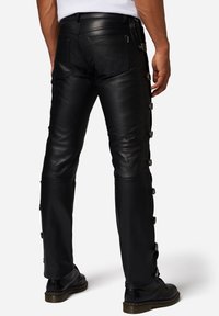 Zwarte leren broek met een strakke pasvorm, zijknopen en ritsdetails. De stof heeft een gladde textuur met een lichte glans.