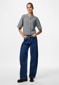 Pilka megzta kardiganas su trumpomis rankovėmis ir mygtukų uždarymu, derinamas su laisvais tamsiai mėlynais džinsais ir juodais batais.