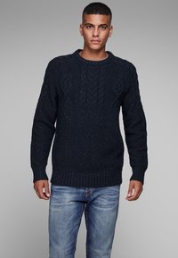 Marineblauwe gebreide trui met een gestructureerd patroon en geribbelde zoom. Bevat kabelgebreide details, gecombineerd met blauwe spijkerbroek.