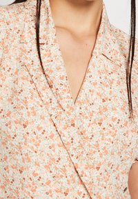 Blouse fleurie avec un motif pêche clair et crème, dotée d'un col, d'un décolleté en V et de détails à boutons. Tissu doux et léger.