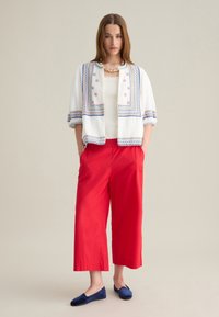 Chaqueta blanca bordada con patrones azules y rojos, combinada con pantalones rojos anchos y zapatos de slip-on azules. Textura de tela suave y ligera.