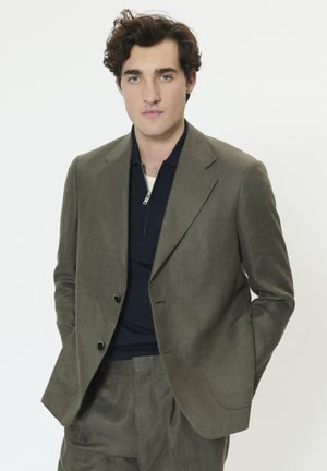 Mladý muž s kudrnatými hnědými vlasy nosí olivově zelený oblek a tmavě modrou košili na zip, stojí před jednobarevným bílým pozadím.