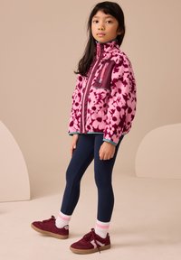 Rózsaszín polárdzseki virágmintával, lila részletekkel, magas gallérral és cipzáros zsebekkel. Tengerészkék leggingssel és bordó sportcipővel párosítva.