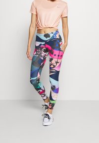 Nike T-shirt in licht perzik met een knoop aan de voorkant, gecombineerd met meerkleurige legging met grafische bloemen- en abstracte ontwerpen. Witte sneakers.