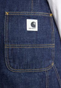 Delle salopette in denim caratterizzate da un colore blu scuro, cuciture dorate e un'etichetta bianca rettangolare con un logo sulla tasca posteriore.