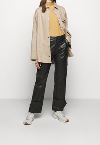 Beige übergroße Hemdjacke, gerippter senfgelber Pullover, schwarze Leder-Hose mit weitem Bein und Cargo-Taschen, weiße Sneakers mit sandfarbenen Akzenten.