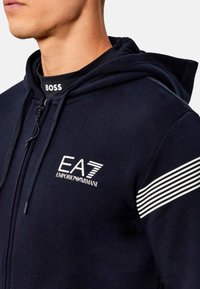 Felpa con zip blu navy con loghi EA7 ed Emporio Armani. Presenta strisce orizzontali bianche sulle spalle e cappuccio con coulisse. Texture morbida.